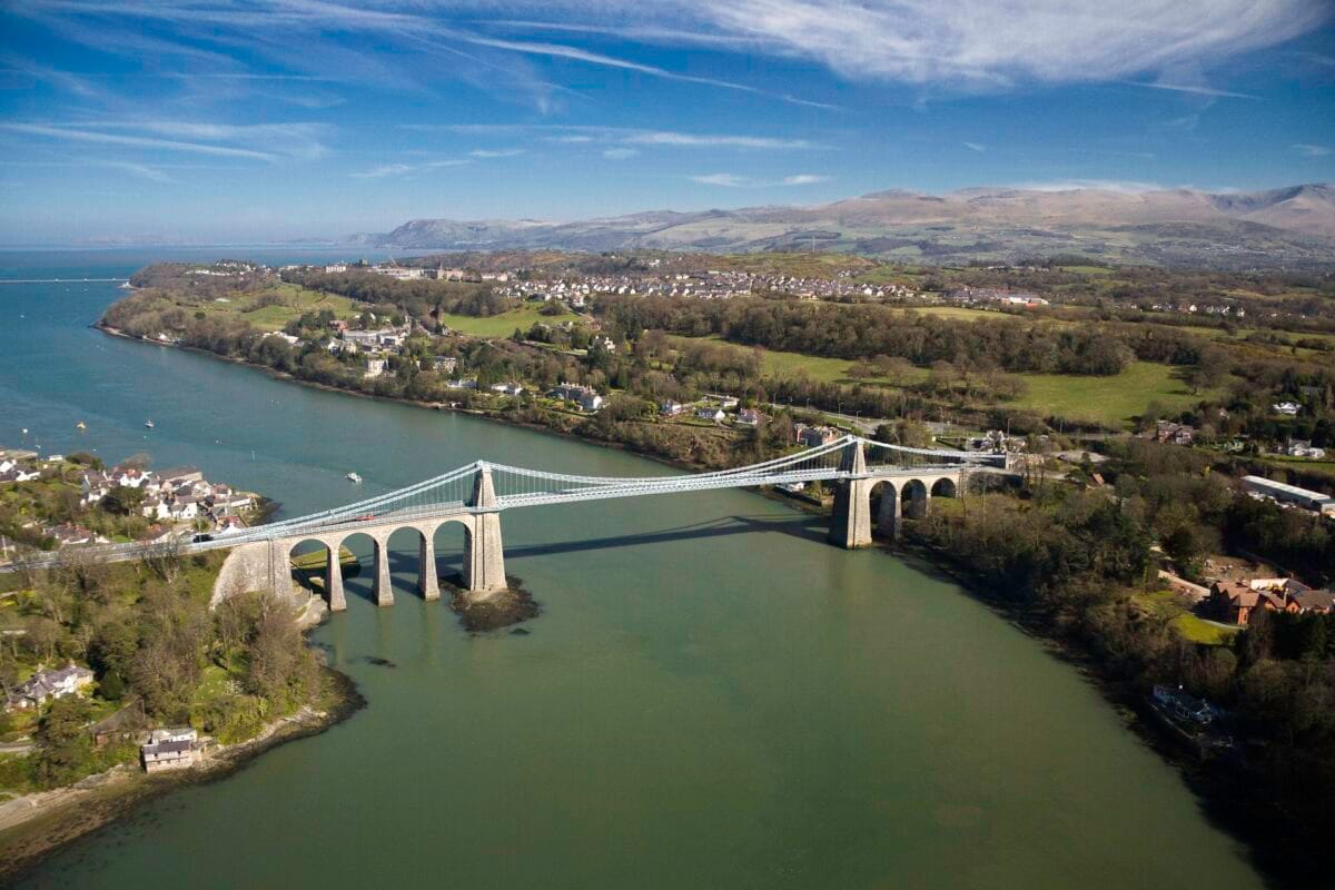 An aerial view of the historic Menai Suspension Bridge stretching across the green waters of the Menai Strait on a bright, sunny day. The bridge features tall stone arches at either end, with lush green coastlines, small town buildings, and the distant mountains of Eryri (Snowdonia) rolling into the background under a blue sky with wispy clouds.