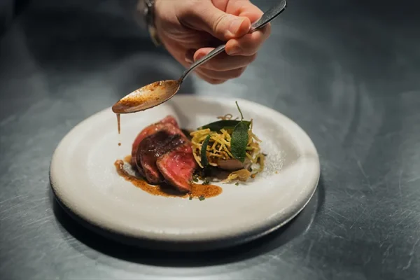 A hand holds a spoon, drizzling sauce over a gourmet dish with slices of meat and shredded vegetables, plated artfully on white—capturing the elegance found in Michelin star restaurants Wales is known for.