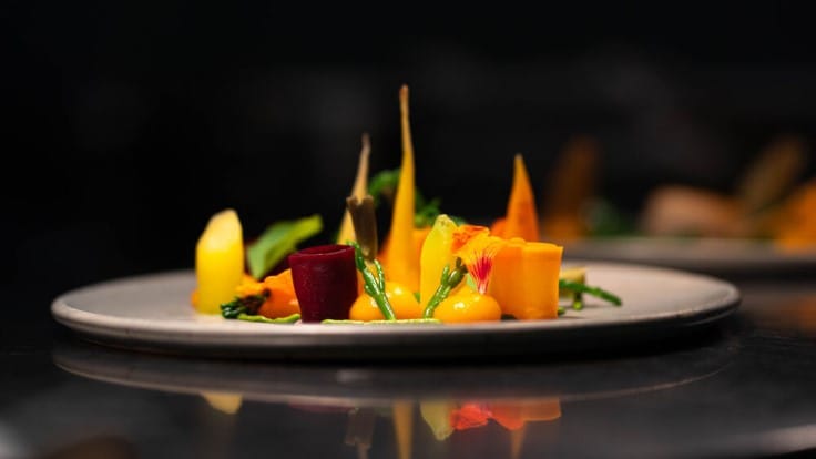 A gourmet dish inspired by Michelin star restaurants Whitebrook in Wales - features artfully arranged colourful carrots and beetroot, garnished with greens and edible flowers, served on a white plate against a dark background.