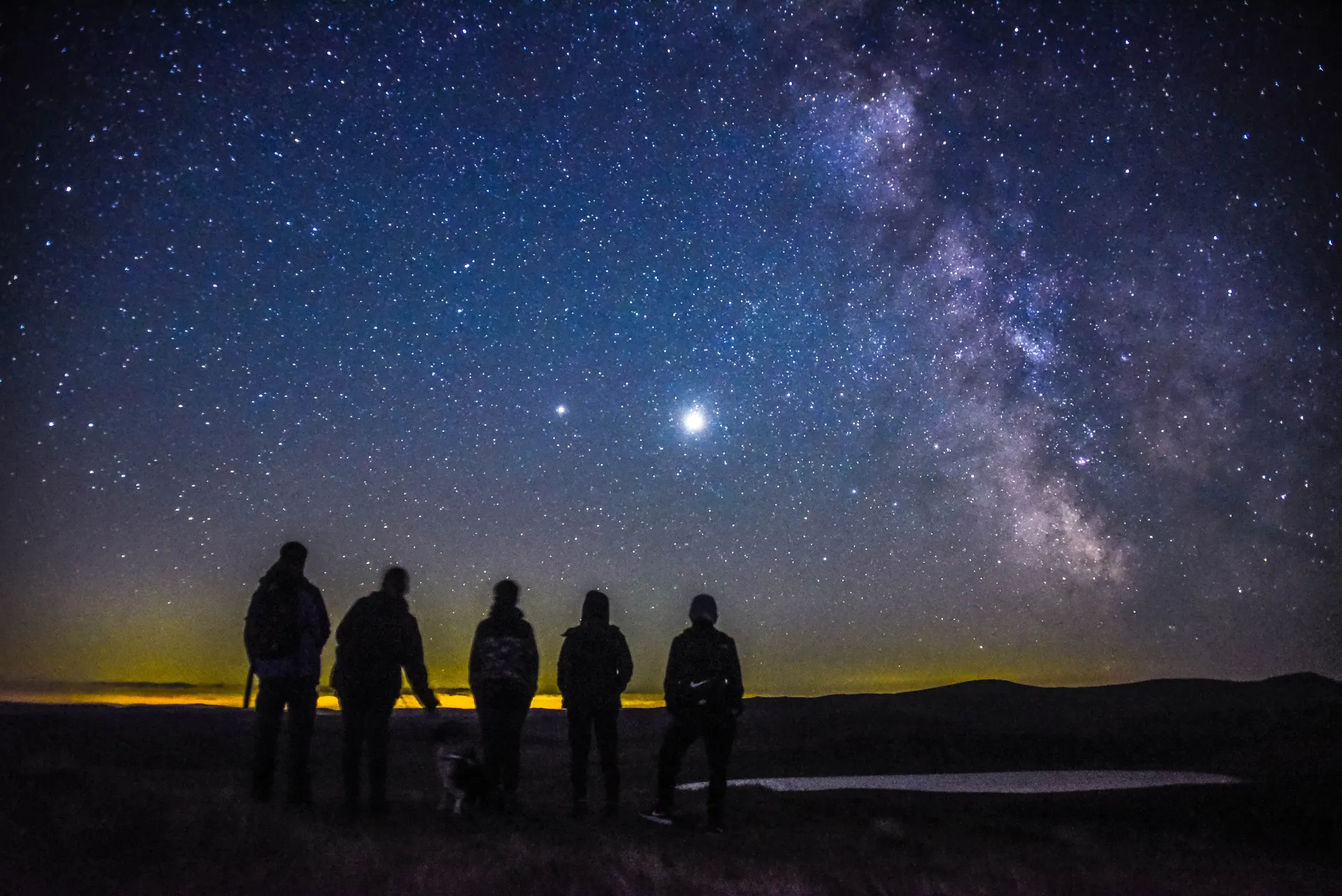 The Milky Way arching across a clear night sky above the silhouetted peaks of Eryri National Park, with the lights of a distant farmhouse visible at the foot of the mountains and stars reflected in still water in the foreground.