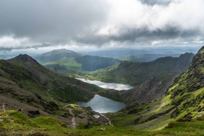 A dramatic landscape of green mountains and valleys with two lakes nestled below, partially illuminated by sunlight breaking through a cloudy sky
