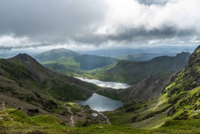 A dramatic landscape of green mountains and valleys with two lakes nestled below, partially illuminated by sunlight breaking through a cloudy sky—perfect for mountain walks in Snowdonia, where tranquillity and rugged peaks surround unspoilt waters.