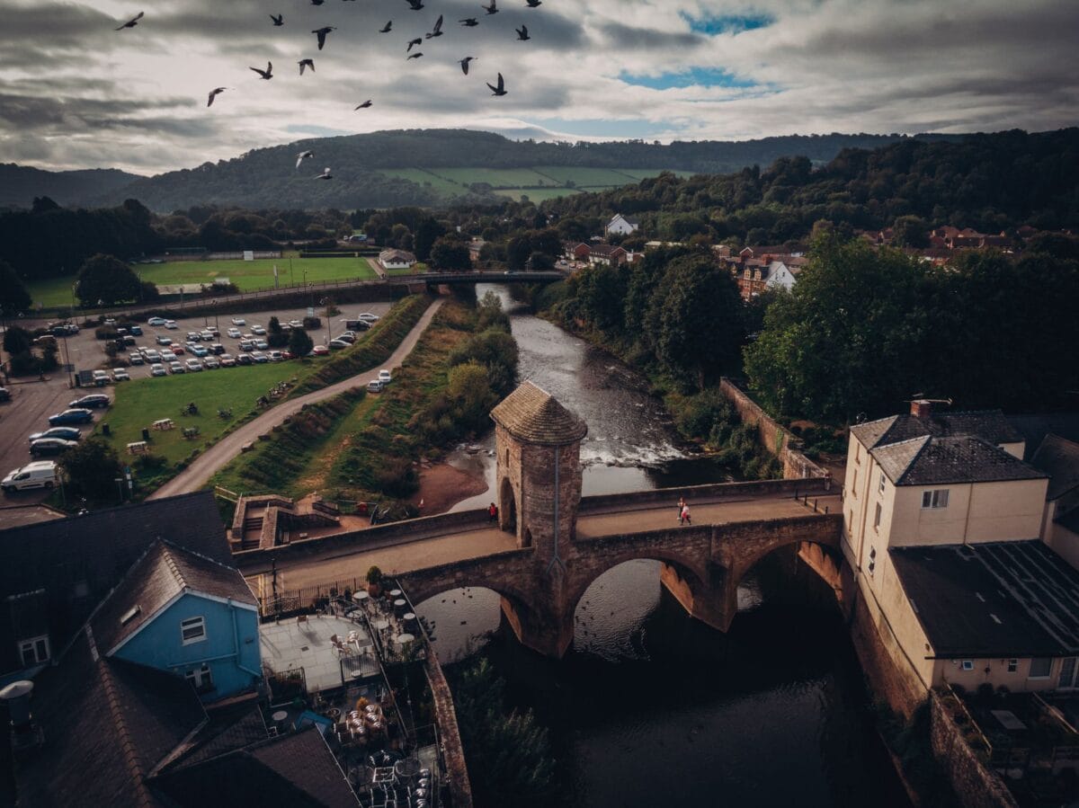 An elevated view of the historic Monnow Bridge in Monmouth, featuring its unique medieval stone gatehouse and arches spanning the dark river, backed by rolling green hills under a cloudy sky.