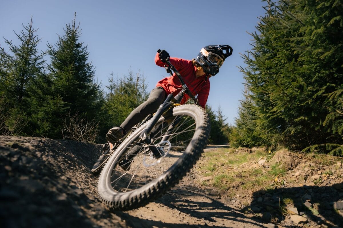 Low-angle view of a rider in a red jersey and full-face helmet leaning hard into a dirt berm while mountain biking North Wales woodland trails under a clear blue sky.