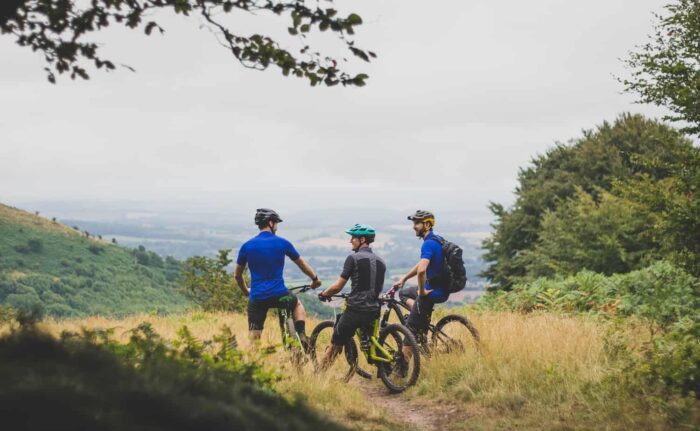 Three cyclists wearing helmets and sports clothing pause with their bikes on a grassy trail, exploring cycling routes in Wales and overlooking a scenic, hilly landscape filled with trees under a cloudy sky.
