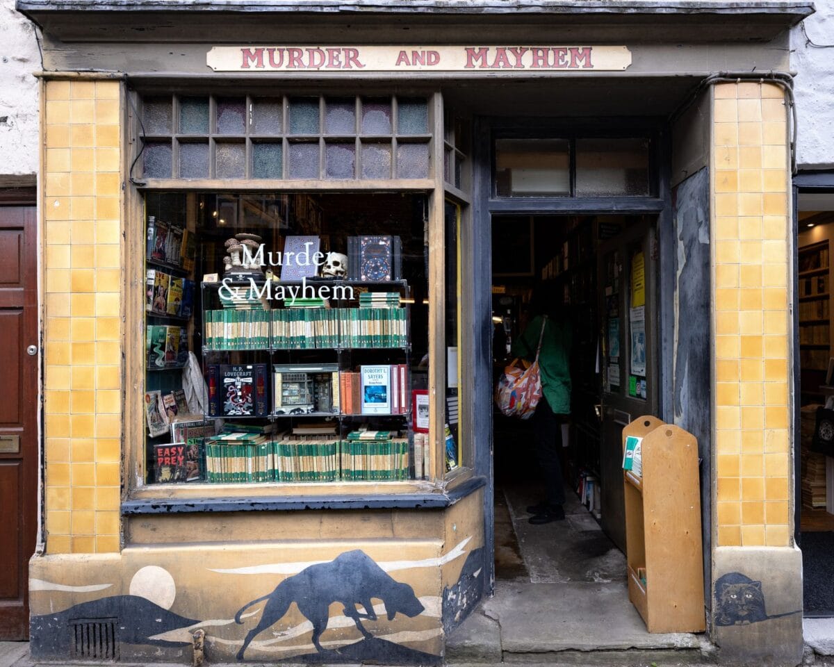 A street-level view of a the bookshop in Haye-On-Wye named 'Murder and Mayhem'. The shop has a yellow tiled exterior and an open doorway. The large glass window displays neat stacks of green-spined mystery books and a decorative skull. Below the window is a painted mural of a large black dog walking across a moonlit landscape.