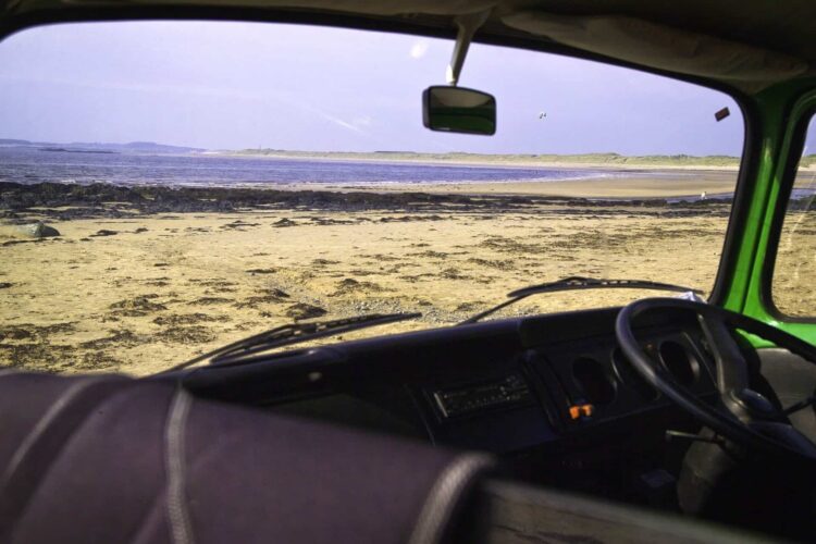 View of a sandy beach and distant water seen through the windscreen of a parked green Wales campervan, with the dashboard and steering wheel visible in the foreground—a perfect snapshot from the Coastal Way Wales.