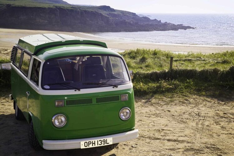 A green vintage camper van is parked on sandy ground near grassy dunes, overlooking a rocky coastline and calm sea—perfect for campervan holidays in Wales along the scenic Cambrian Way in Wales under a clear sky.