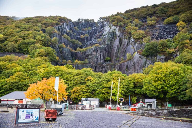A scenic view of the National Slate Museum in Llanberis features steep rock faces, dense green trees, signs, railway tracks, a bright tree with yellow leaves, and small buildings under an overcast sky.