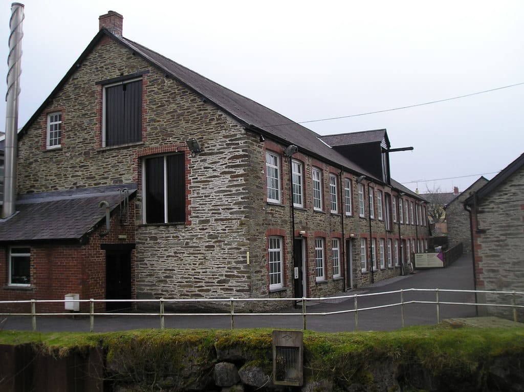 The exterior of a long stone and brick mill building with slate roofs and a chimney, part of the former Cambrian Mills complex, viewed across a moss-covered mill pond wall.