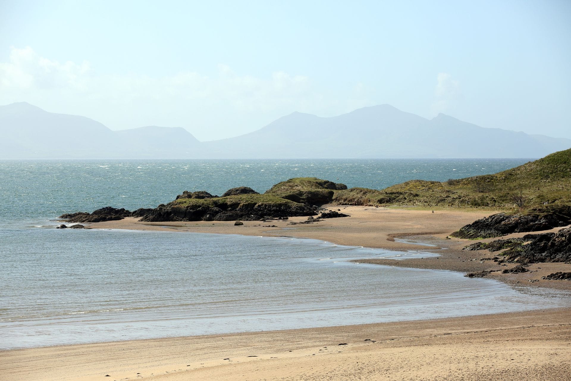 Newborough Beach on Anglesey at low tide, with the headland of Ynys Llanddwyn visible as a green peninsula extending into the Irish Sea and the snow-capped Eryri mountains in the background.