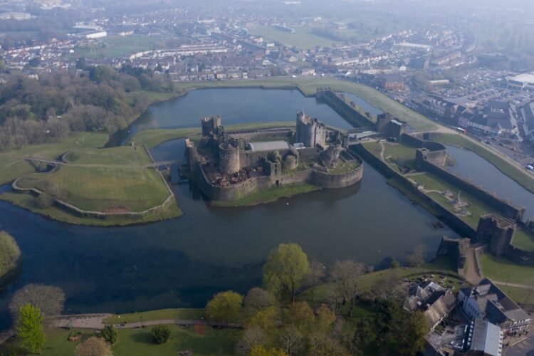 An aerial view of the impressive Caerphilly Castle, featuring a massive stone fortress completely surrounded by large, reflective water moats and vibrant green earthwork defenses. Stone bridges connect the central stronghold to the outer grounds, while a sprawling town with rows of houses and streets fills the distant background under a slightly hazy sky.