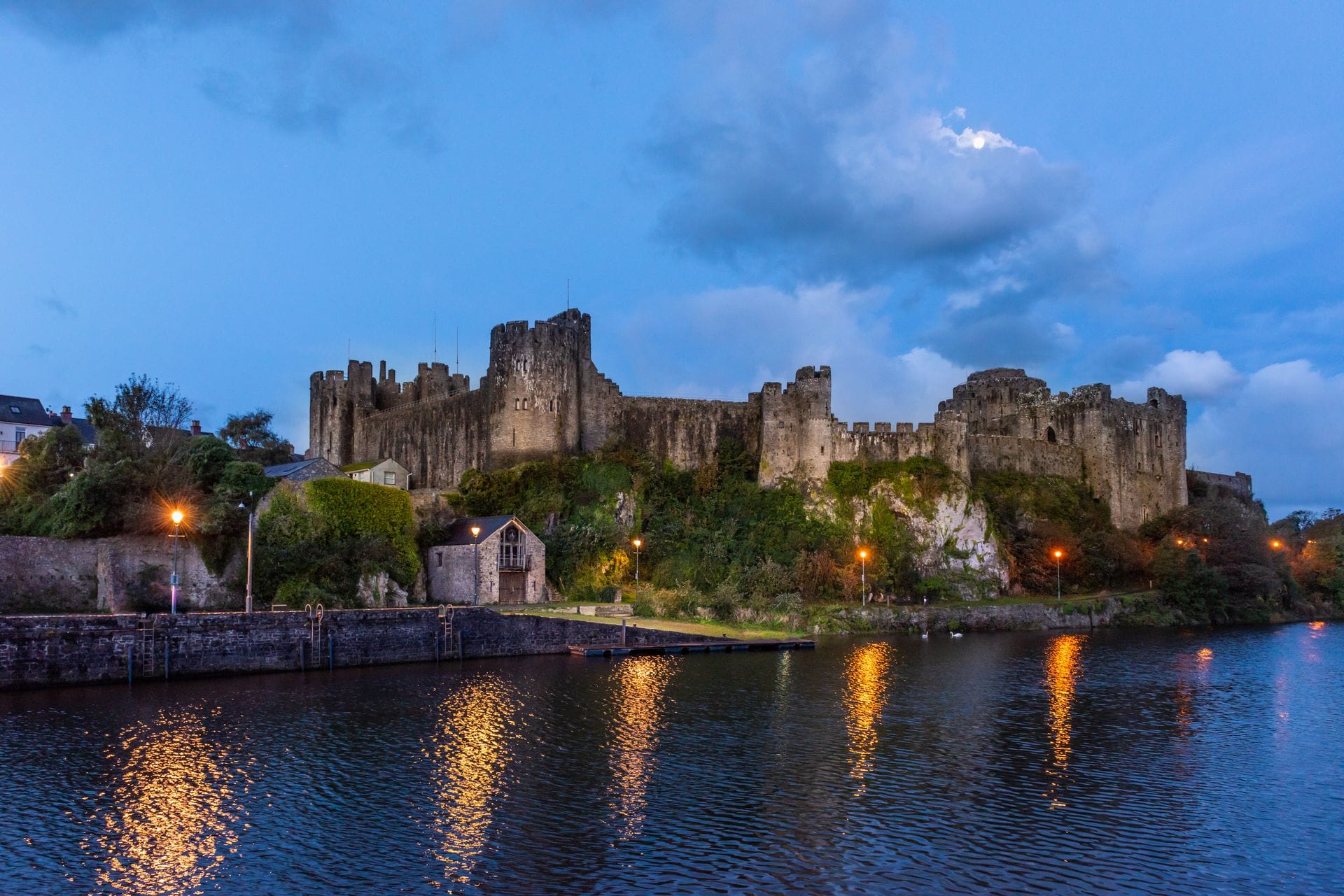 Pembroke Castle illuminated at dusk, its towers lit against a deep blue sky with the moon visible above, reflected in the still water of the Mill Pond below.