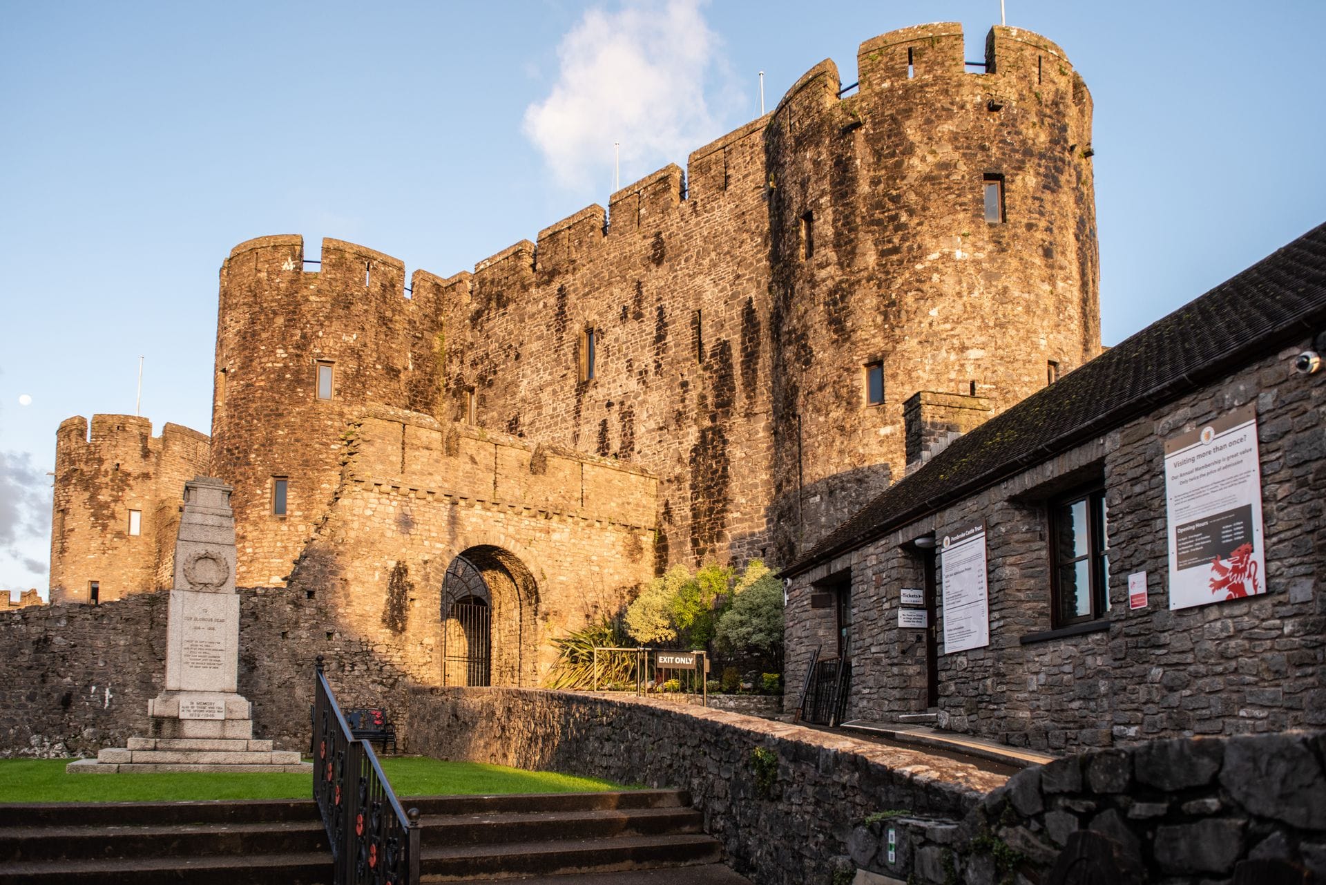 The entrance to Pembroke Castle at ground level, showing round towers and crenellated walls beside the visitor centre and ticket office, with a war memorial in the foreground.