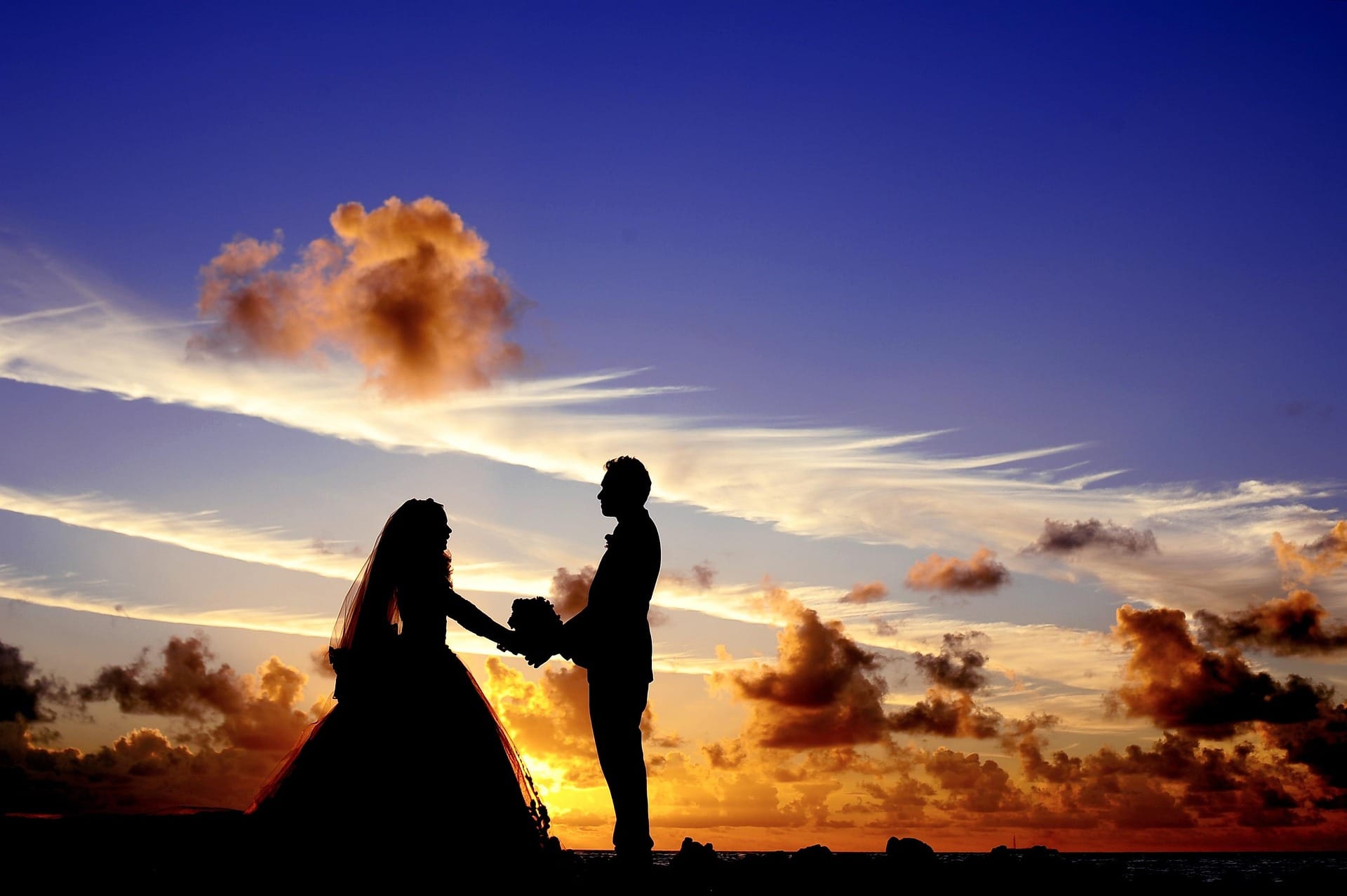 A silhouette of a bride in a full gown and veil standing face-to-face with a groom, holding hands against a spectacular, dramatic sunset sky filled with glowing golden and dark orange clouds.