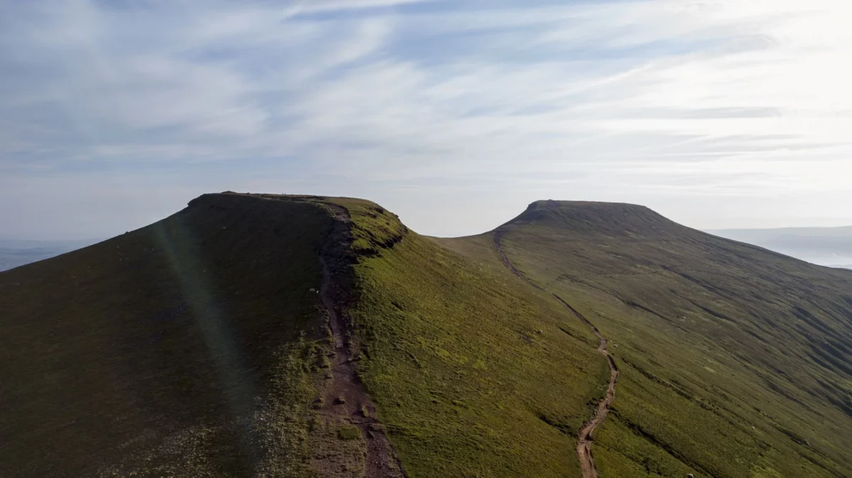 A worn dirt hiking trail winds along a steep, grassy ridge connecting two flat-topped mountain peaks at Pen y Fan in Mid Wales.