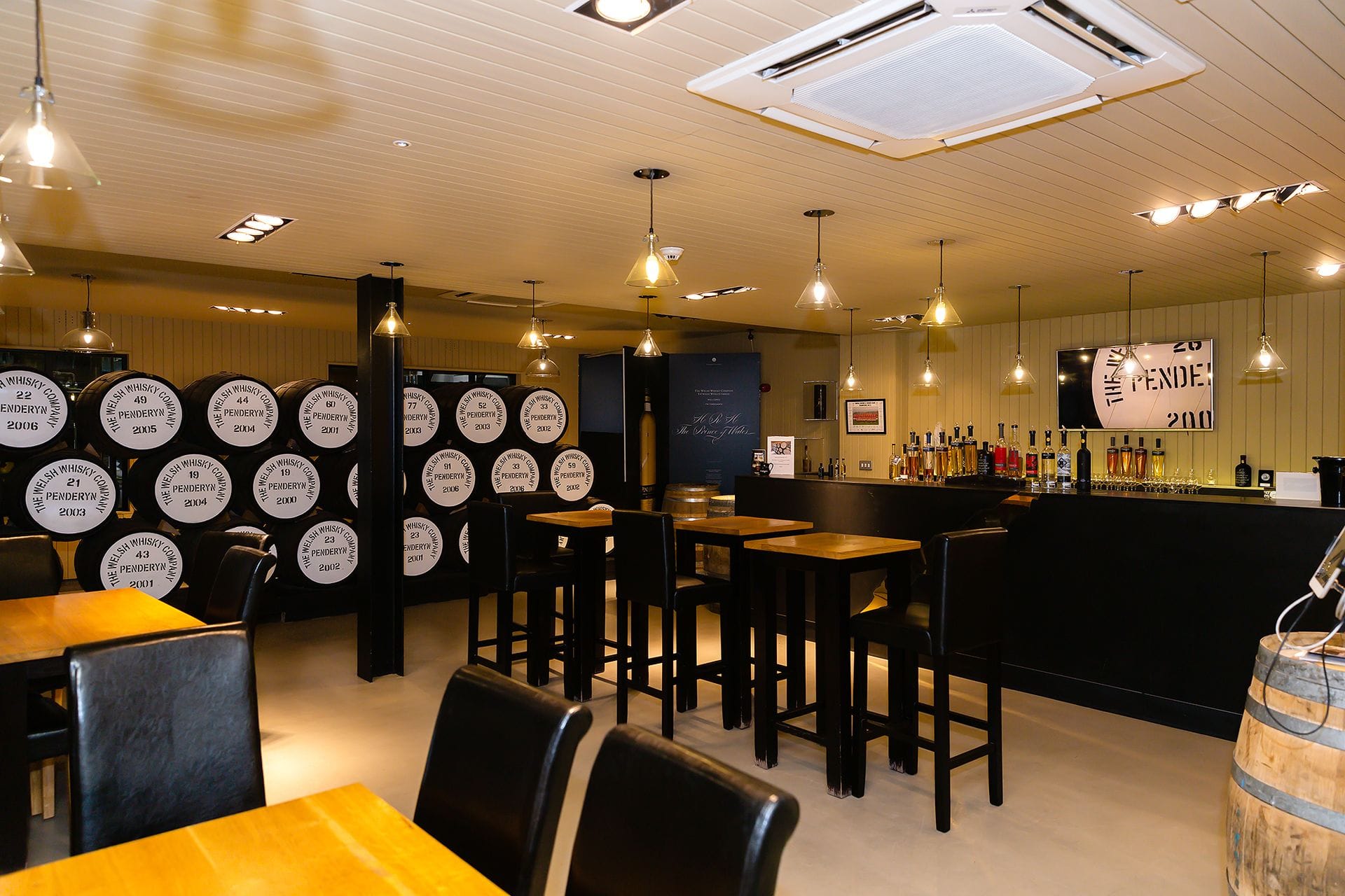 The modern tasting room and visitor bar at Penderyn Distillery in Wales, featuring wooden tables, seating areas, and a prominent wall displaying stacked wooden whisky casks.