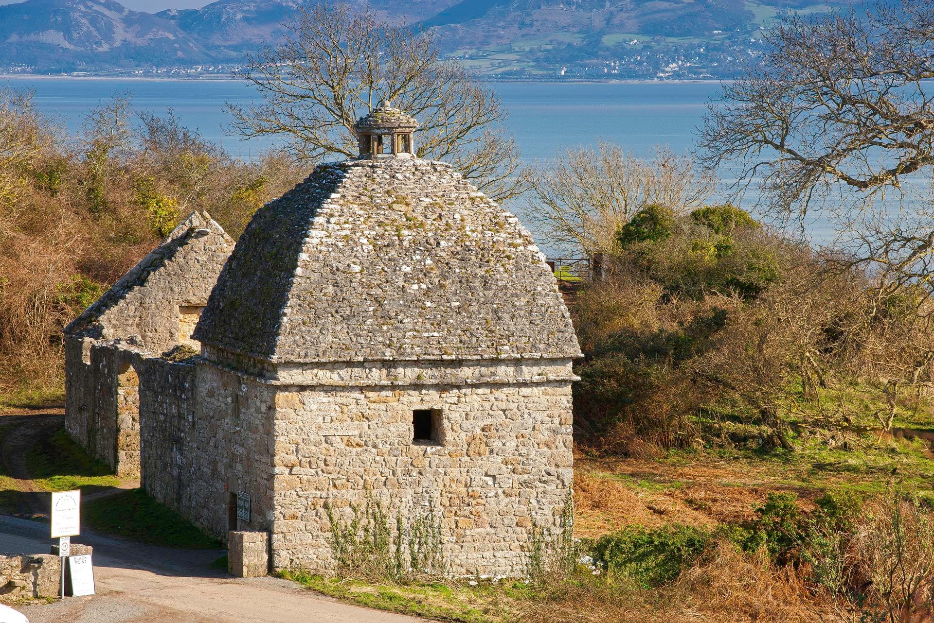The distinctive round stone dovecote at Penmon, with its conical slate roof and small lantern tower, set beside a country lane with the Menai Strait and mountains visible in the background