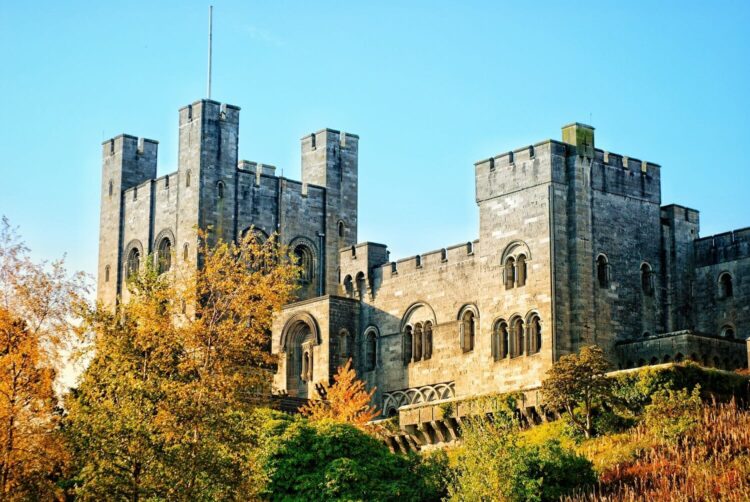 Penrhyn castle with tall towers and arched windows, surrounded by colourful autumn trees under a clear blue sky.