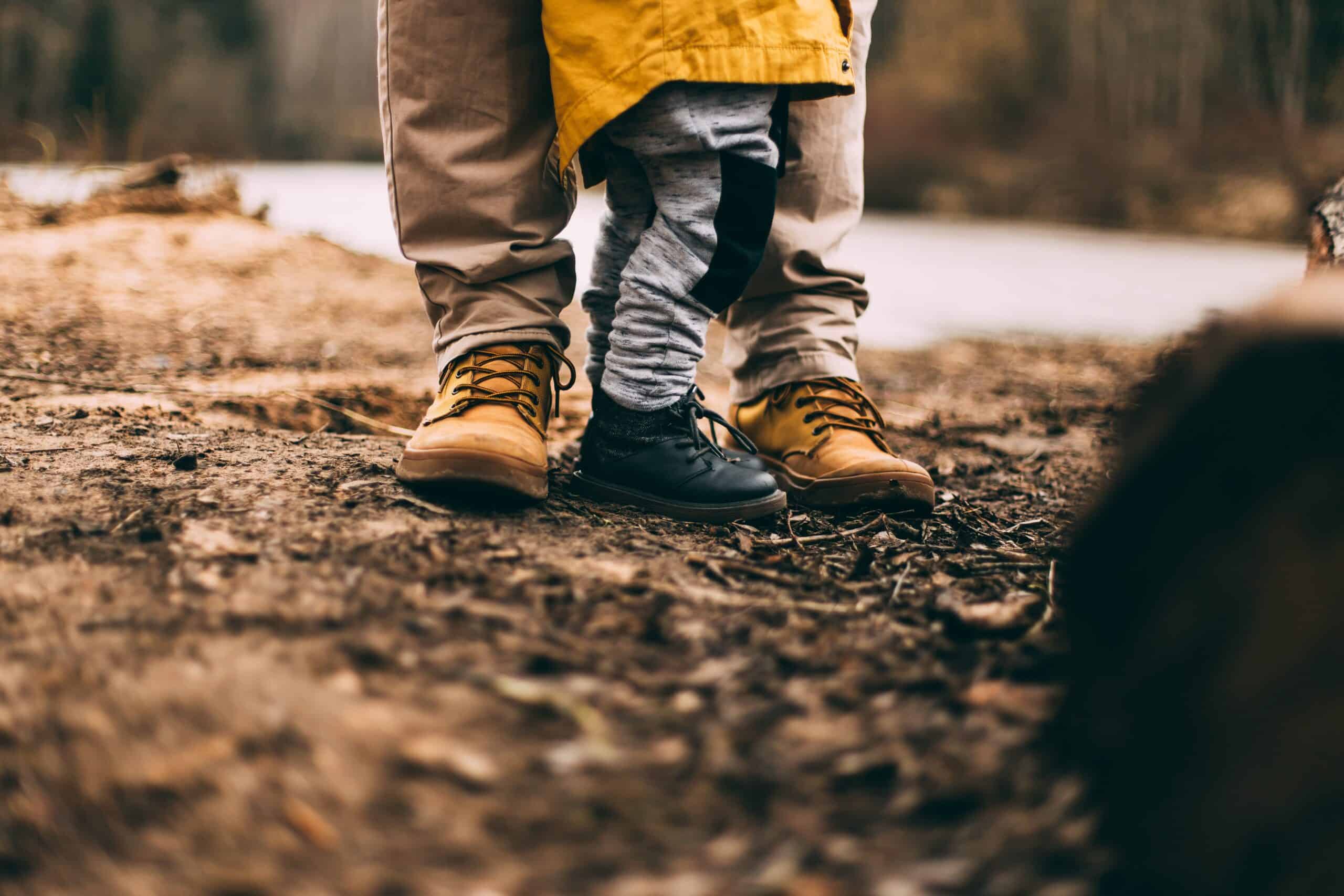 A close-up, ground-level photograph of an adult's and young child's feet on a woodland trail near a body of water. The adult wears sturdy brown hiking boots, and the small child stands snugly between their legs wearing dark boots, patterned trousers, and the edge of a yellow waterproof jacket. The ground is covered in muddy dirt and autumn debris.