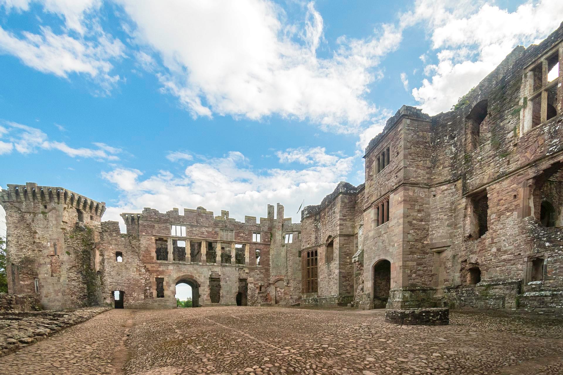 The cobbled Pitched Stone Court at Raglan Castle, surrounded by ruined multi-storey stone ranges with large empty window openings, under a partly cloudy sky.