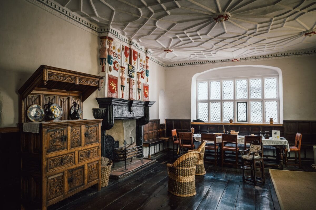 An atmospheric Elizabethan room inside Plas Mawr, featuring an intricately patterned white plasterwork ceiling, a large fireplace adorned with brightly painted heraldic crests, dark wooden floorboards, and antique carved wooden furniture.