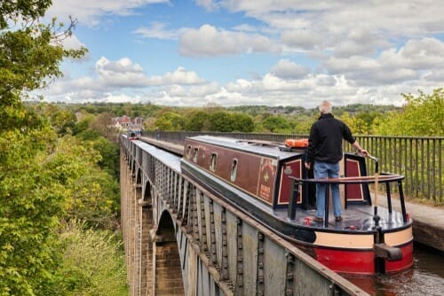 A man standing at the back of a traditional narrowboat, steering it across the impressive, high Pontcysyllte Aqueduct. The narrow metal structure carries the canal water high above a lush green valley filled with trees, with rolling hills in the background under a bright sky dotted with fluffy white clouds.