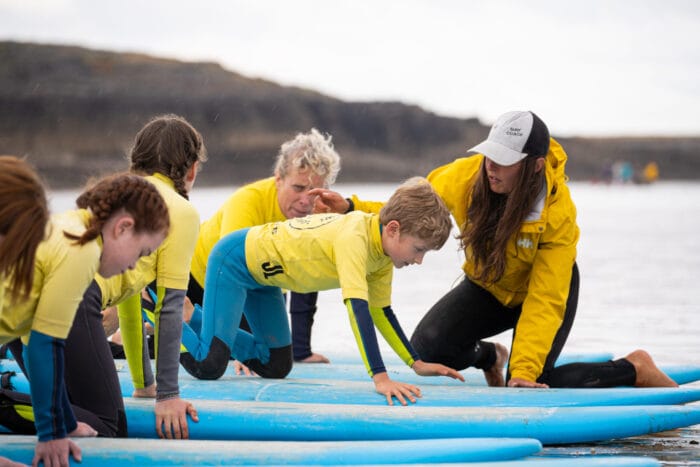 A group of children in yellow shirts practise surfing moves on blue boards at one of the best surfing beaches in Wales, guided by an adult instructor in a yellow jacket and white hat. The scene is set near the water, with rocky cliffs in the background.