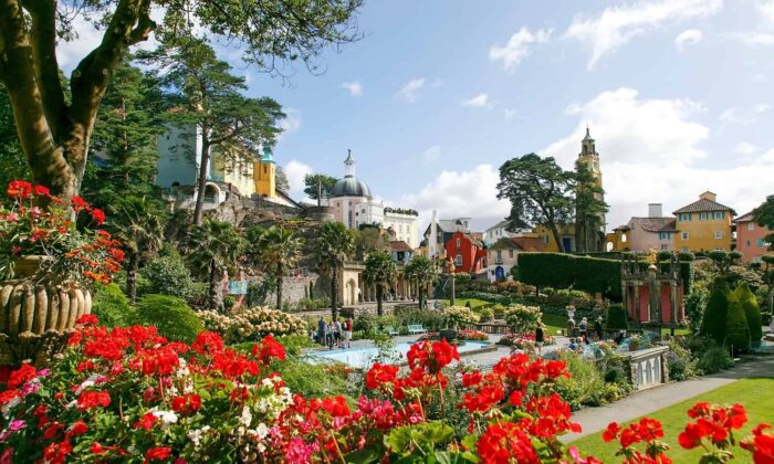 Colourful buildings and lush gardens filled with red flowers and greenery under a partly cloudy sky make Portmeirion one of the most beautiful places in Wales, creating a vibrant, picturesque village scene.