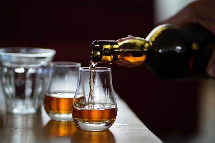 A hand pours amber-coloured whisky from a bottle into a glass on a wooden table, with another whisky glass and a water glass in the background—echoing the craftsmanship found at distilleries in Wales.