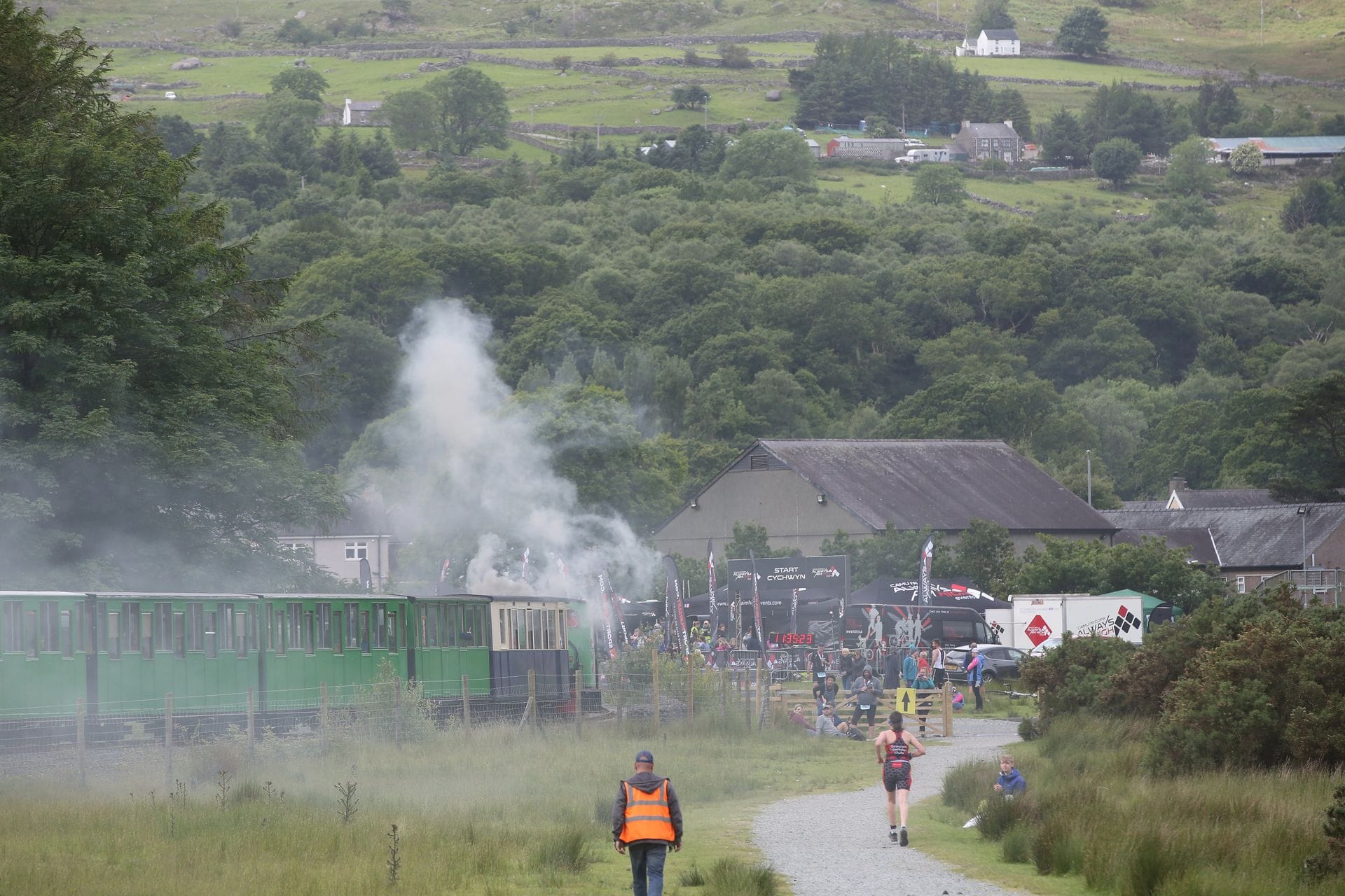 A runner on a gravel path races alongside a vintage green steam train billowing white smoke through the lush countryside. Event tents and a crowd of spectators are gathered in the background, highlighting one of the most unique sports events in Wales.