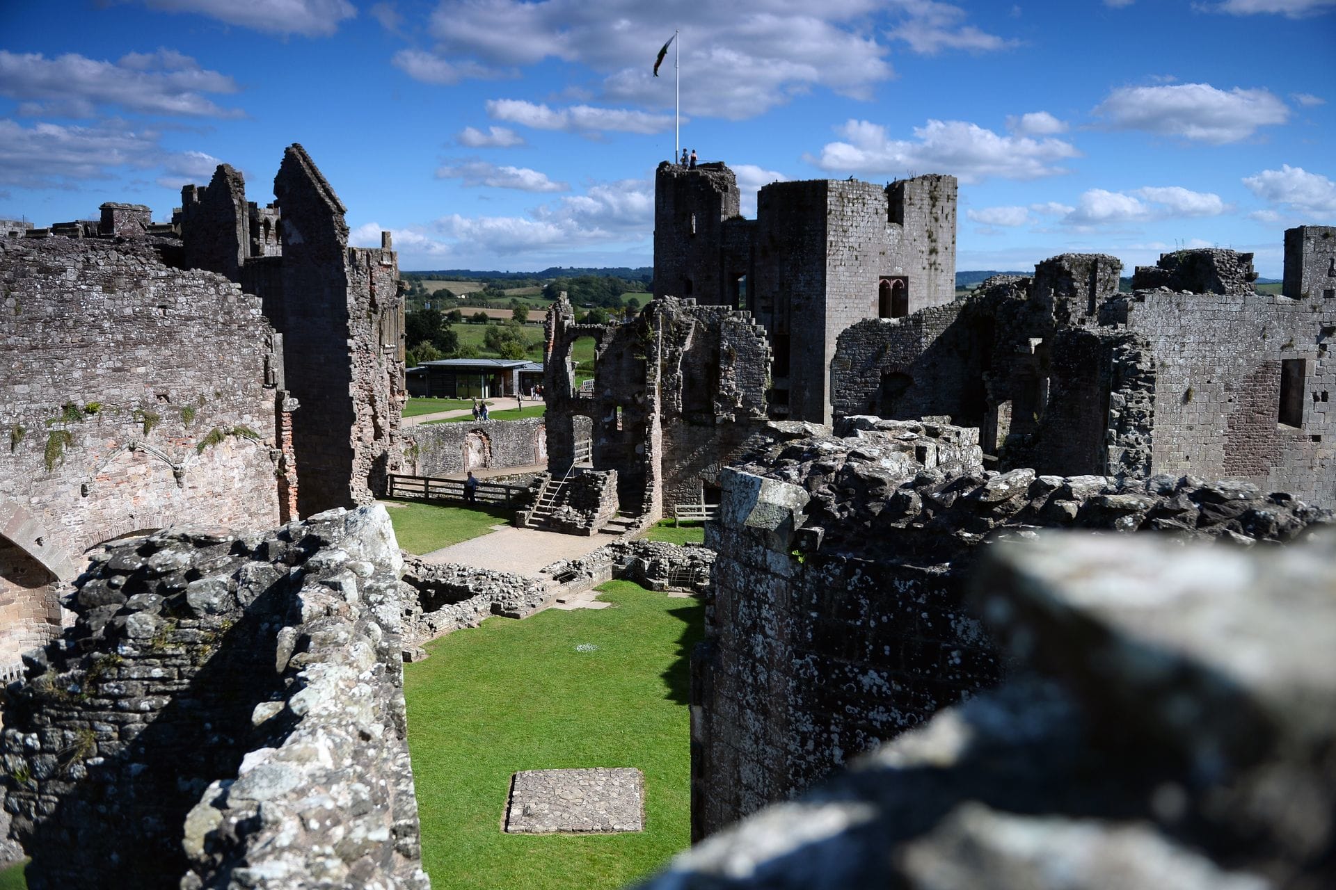 An elevated view through Raglan Castle's ruined walls across a grassed inner courtyard, with visitors standing atop a tower flying the Welsh flag and rolling Monmouthshire countryside visible in the distance.