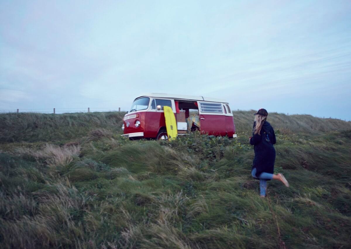 A person wearing a dark coat and cap walking up a rugged, grassy hill towards a vintage red and white campervan. The van's side door is open, and a bright yellow surfboard is resting against it under an overcast evening sky.