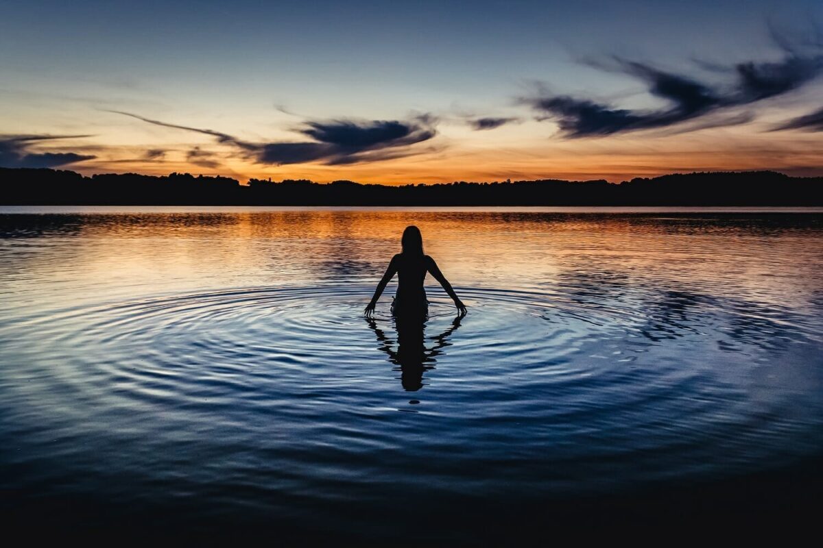 Silhouette of a person wild swimming in a calm lake in Wales, surrounded by gentle ripples reflecting a vibrant orange and blue sunset.