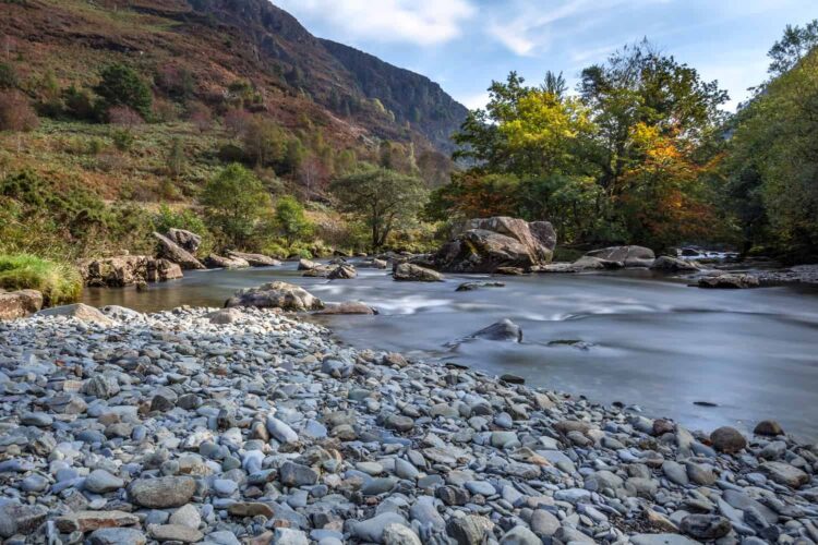 The river Glasyn flows over smooth rocks with a pebbled shore in the foreground, surrounded by lush trees and hills—an idyllic scene reminiscent of mountain walks in Snowdonia near Porthmadog under a partly cloudy sky.