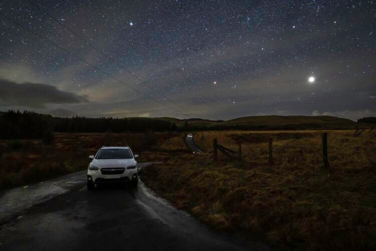 A white car is parked on a narrow country road at night, grassy fields on either side and rolling hills ahead—a tranquil scene for anyone seeking Wales road trip ideas beneath a star-filled sky, with the moon or a bright planet shining above the horizon.