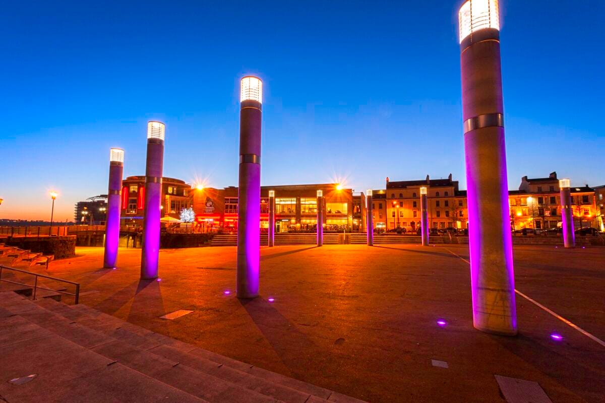 A twilight view of the Roald Dahl Plass public square in Cardiff Bay. Tall stone pillars are lit with bright purple lights against a deep blue evening sky, with warmly lit restaurants in the background. Visiting this waterfront spot is one of the best things to do in Cardiff.