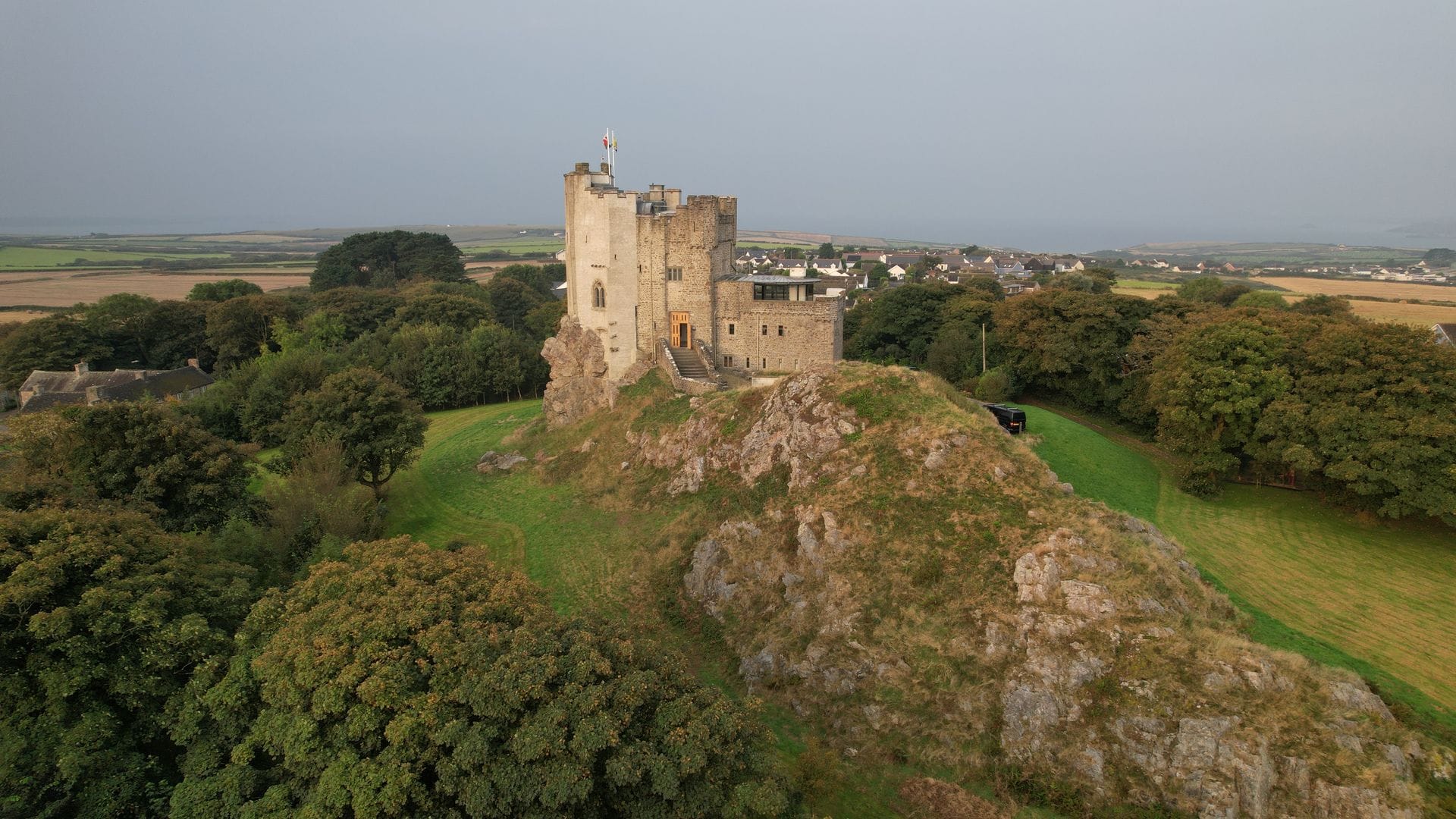Aerial view of Roch Castle, a pale stone tower with flags on top and a modern glass-walled extension, standing on a rocky, grass-covered outcrop among trees, with fields, village houses and the sea beyond.