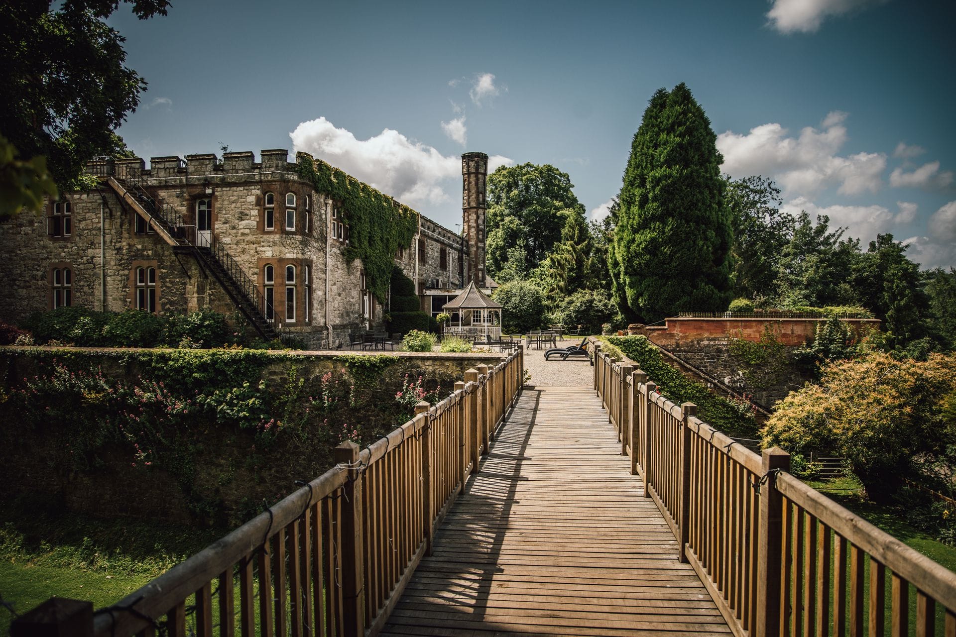 A wooden footbridge leading to a gravel terrace with a gazebo and sun loungers, beside the ivy-covered castellated stone front of Ruthin Castle Hotel, with a tall slender tower behind and a large conifer on the right.