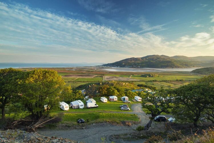 A scenic view of a caravan site in Wales, with several white caravans parked on grassy fields, surrounded by green trees, hills, and mountains under a blue sky with scattered clouds.