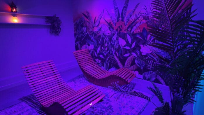 Two wooden loungers in a dimly lit spa relaxation room with purple-blue lighting, tropical wall art, potted plants, and a glowing lantern and candle