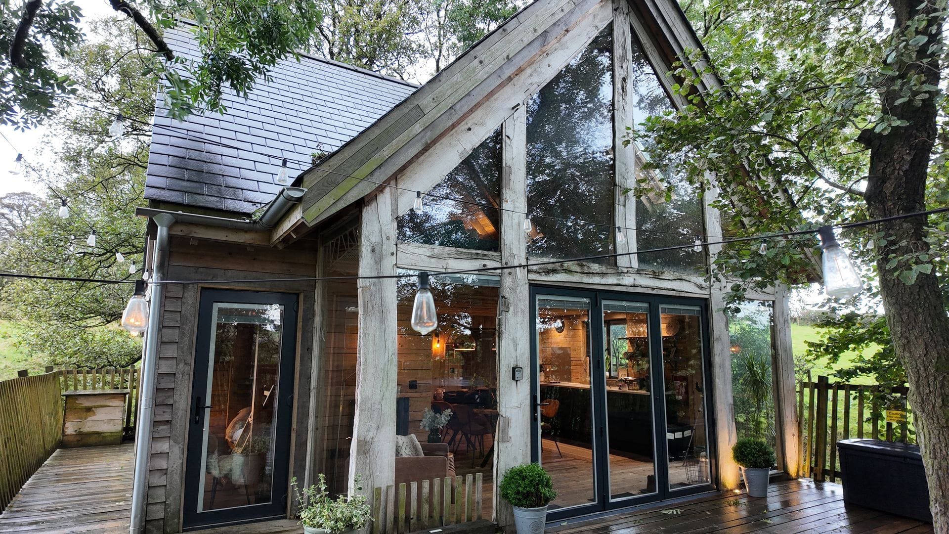 A timber-framed treehouse cabin with a slate roof, large A-frame glass windows, and a wooden decking area surrounded by green trees, with decorative string lights hanging across the exterior.