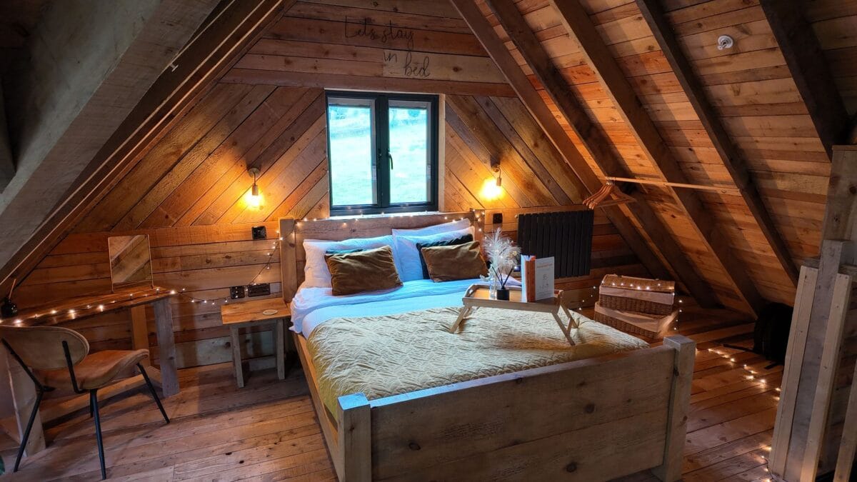 A cosy loft bedroom interior in a wooden cabin, featuring a bed with fairy lights, vaulted ceilings, and a breakfast tray for unique romantic accommodation in Wales.