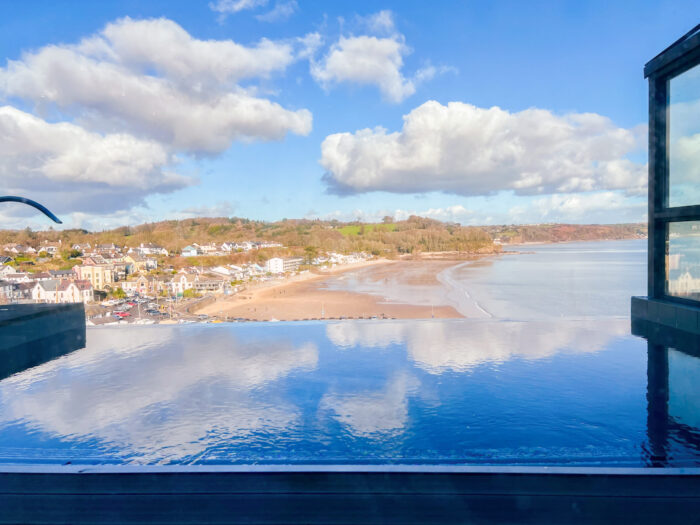 An infinity pool at St Brides Spa Hotel in Wales overlooks a sandy beach and a coastal town with houses and trees under a blue sky with scattered clouds.