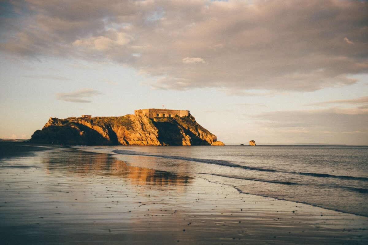 A scenic sunset view of the historic St Catherine's Island fort sitting on a rugged rocky outcrop, viewed across the calm, low-tide shoreline of Tenby
