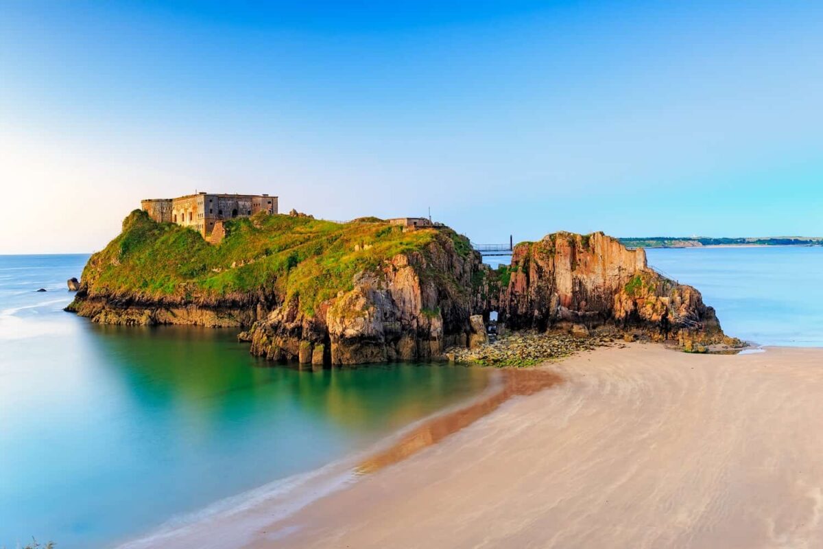 A wide coastal shot showing a large, grass-topped rocky island featuring an old stone fort. The island is surrounded by calm turquoise water and a wide stretch of golden sand accessible during low tide under a clear blue sky.