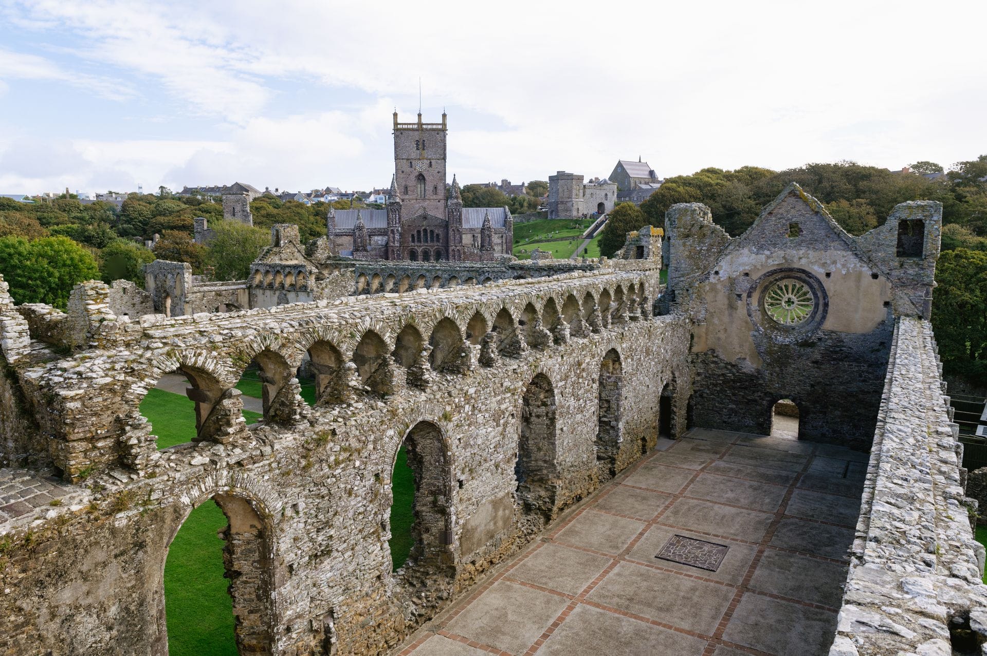 The medieval ruins of the Bishop's Palace at St Davids, showing the surviving stone arcaded parapet, great hall walls, and ruined chambers, with St Davids Cathedral visible across the River Alun in the background.