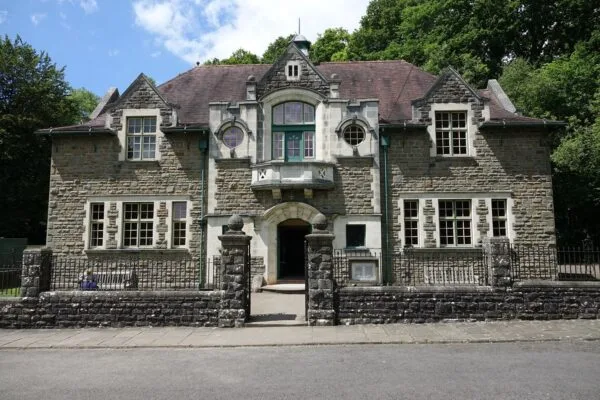 The front of St Fagans Museum features a stone building with a steep, dark red roof, arched entrance, and decorative windows, surrounded by trees and a low stone wall under a partly cloudy sky.