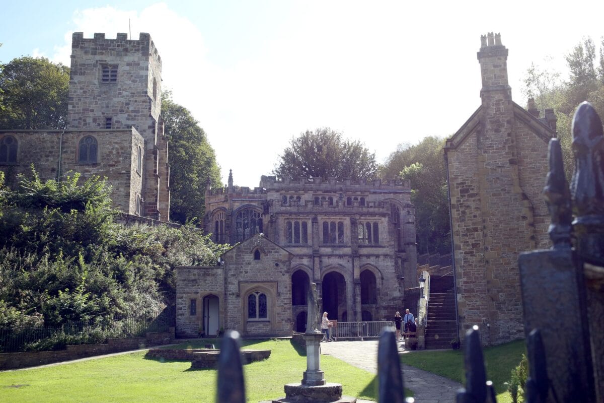 Looking past the blurred spikes of a dark metal fence towards a complex of historic stone buildings. In the centre is the ornate Gothic chapel of St Winefride's Well with large arched entrances, flanked by a square stone church tower on a grassy, tree-lined hill to the left.
