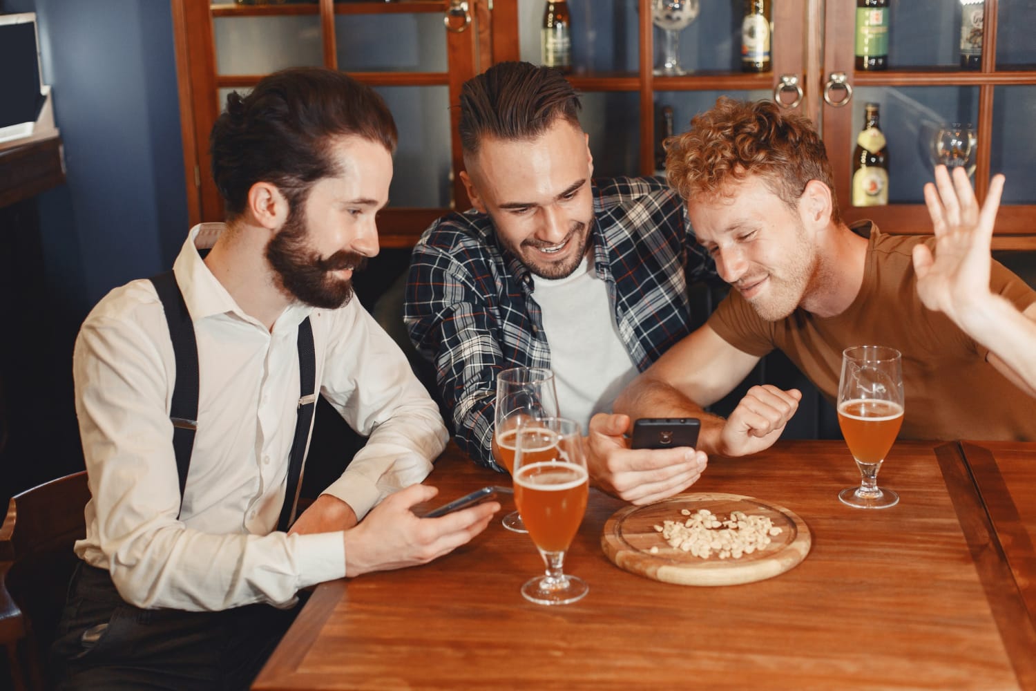 Three men smiling and looking at a smartphone together while sitting at a wooden table with glasses of beer and snacks, enjoying a stag party retreat.